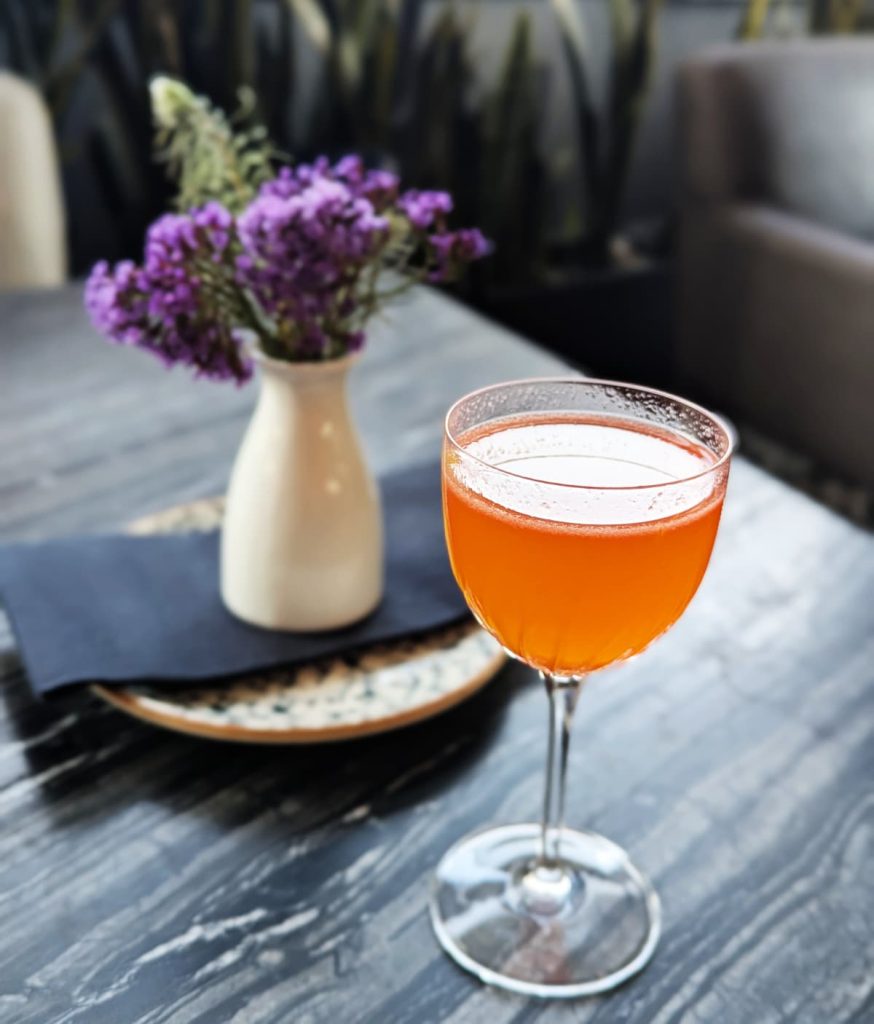 Orange cocktail in a coupe glass on a dark table at a Zócalo restaurant in Mexico City. In the background is a small vase of purple flowers. The drink catches the light and sets the mood for sunset drinks with a view.