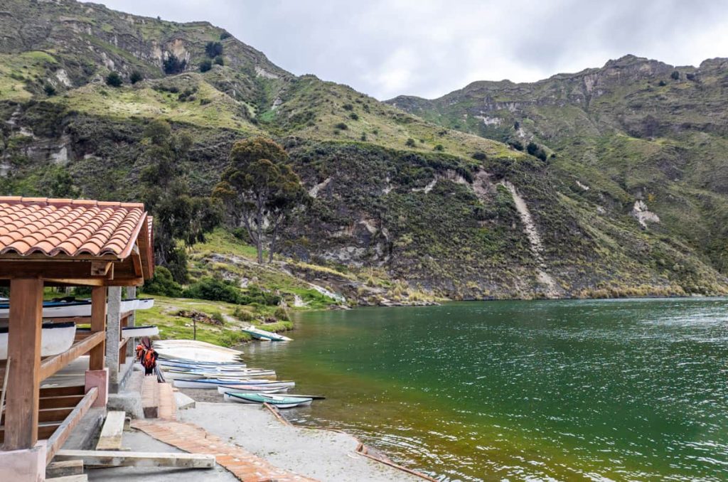 Small rowboats lined up along the edge of Quilotoa Lake beside a red-roofed structure, with steep green hills rising in the background. Many visitors choose to kayak in Laguna de Quilotoa.
