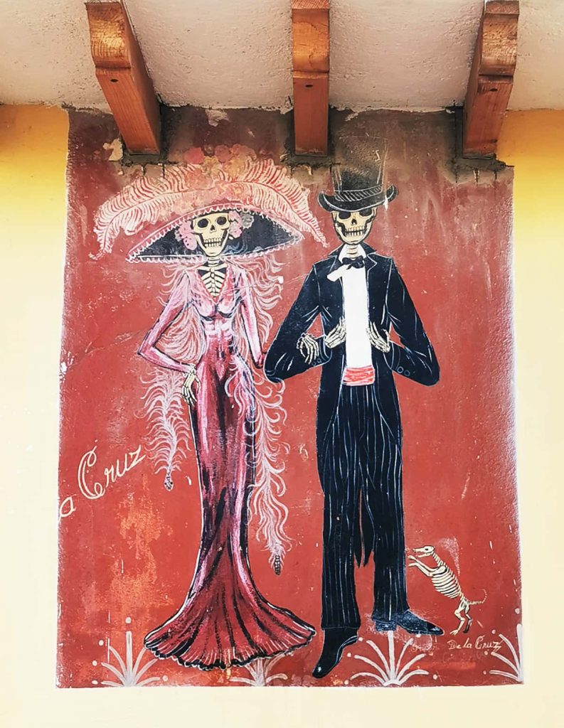 Street mural with a red background on a yellow wall depicting La Catrina in a pink gown, wide hat, and feather boa alongside El Catrín in a black suit and top hat, with a small skeleton dog at his feet.