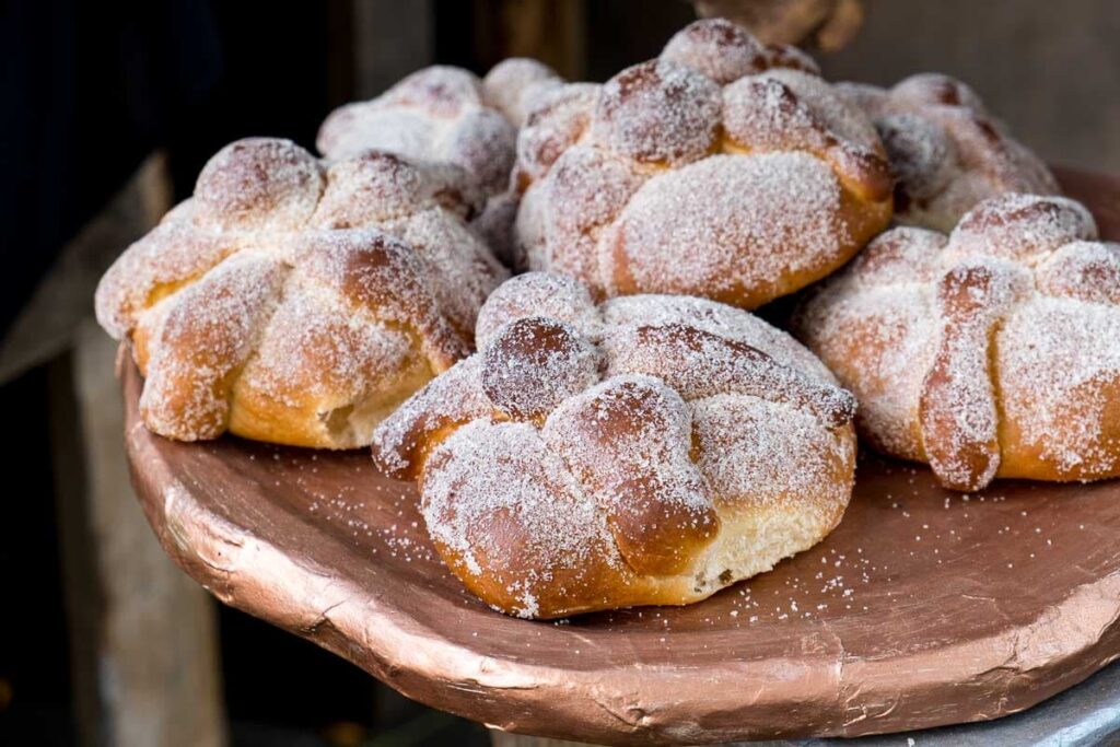 A close-up of a clay plate six pieces of pan de muerto, one of the most traditional Day of the Dead foods. Each is in the shape of bones and dusted with sugar.