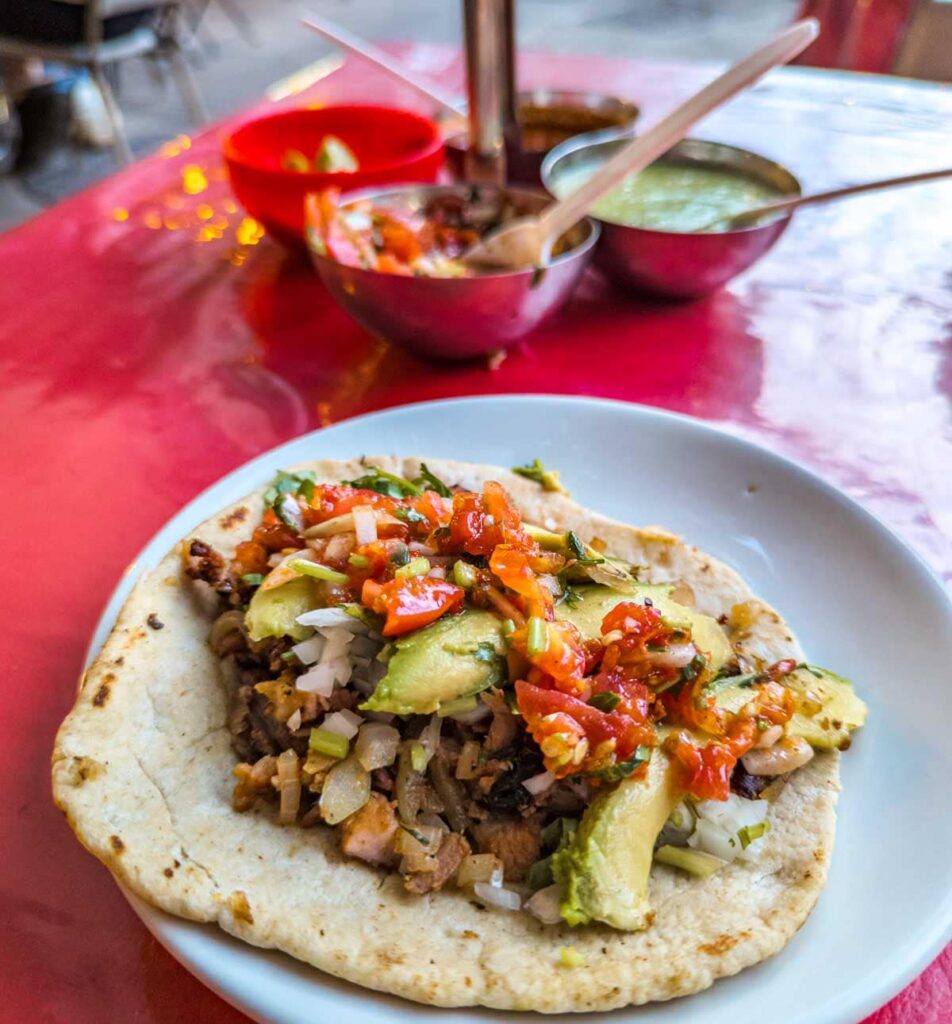 Taco arable in Mexico City on a white plate sits on a red tablecloth with three small bowls of salsa in the background and a bowl of limes.